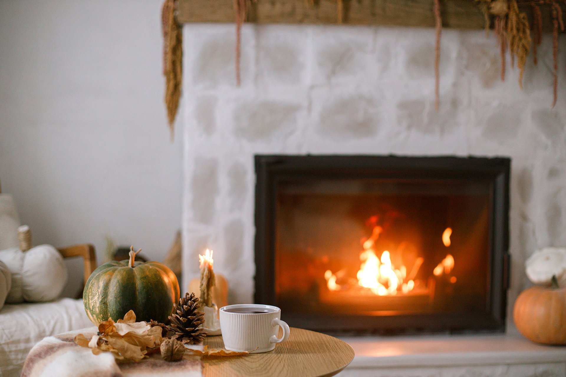 Gemütliche Wohnzimmerszene mit brennendem Kamin, herbstlicher Dekoration aus Kürbissen, Kiefernzapfen, getrockneten Blättern und einer Tasse Kaffee auf einem Holztisch, vermittelt ein Gefühl von Wärme und Geborgenheit im Zuhause.