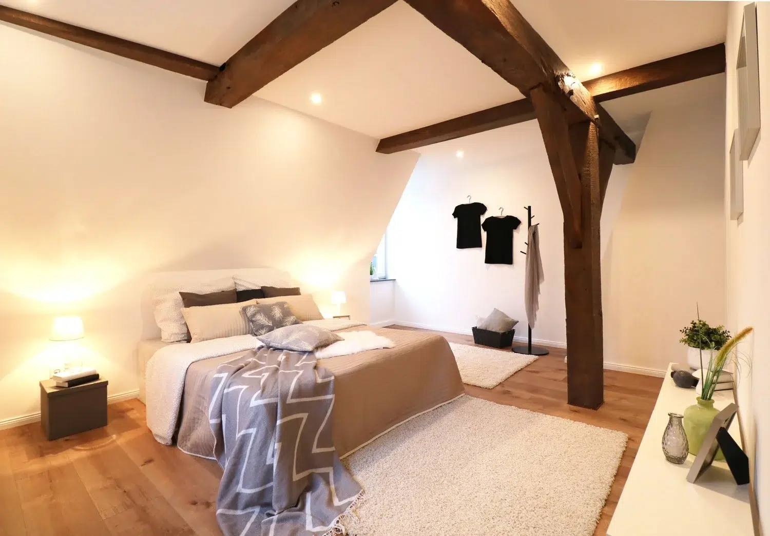 Modern eingerichtetes Schlafzimmer mit Holzbalken, Holzboden, einem großen Bett mit beige-brauner Bettdecke und mehreren Kissen, weichen Teppichen sowie moderner Dekoration und Beleuchtung in einem hellen Dachgeschoss.