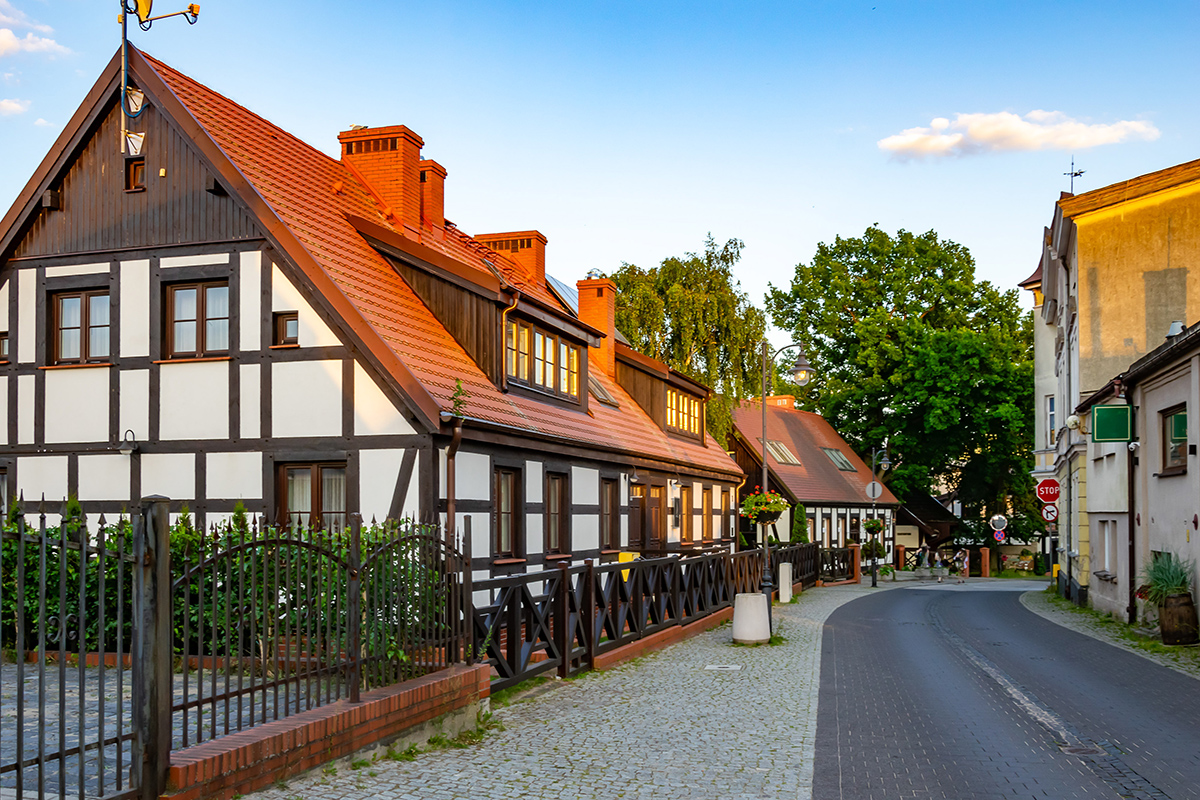 Eine ruhige, kopfsteingepflasterte Straße mit traditionellen Fachwerkhäusern mit roten Ziegeldächern, umgeben von Bäumen und einem blauen Himmel bei Sonnenuntergang.